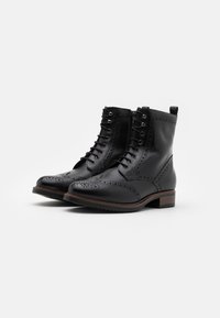 Botas de tobillo de cuero negro con detalles de brogue, cordones en la parte frontal, cremalleras laterales y tacones apilados marrones sobre un fondo blanco.