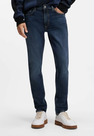Mand iført mørkeblå slim-fit jeans med hvide sneakers mod en ensfarvet baggrund.