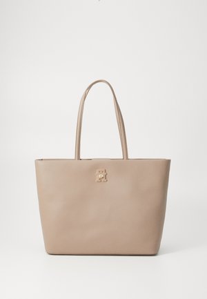 Beige tygväska tillverkad av slät läder, med två långa handtag och en guldfärgad logga på framsidan. Minimalistisk rektangulär design.