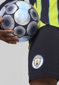 Nogometna žoga v Manchester City slogu z modrimi in srebrnimi paneli, ki prikazuje krožno vzorčenje. Igralec nosi črne hlače s logotipom in zelenimi detajli.