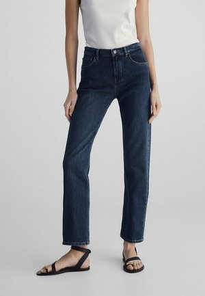 Donna con jeans blu scuro a gamba dritta, top bianco senza maniche e sandali neri con dettaglio dorato su sfondo semplice.