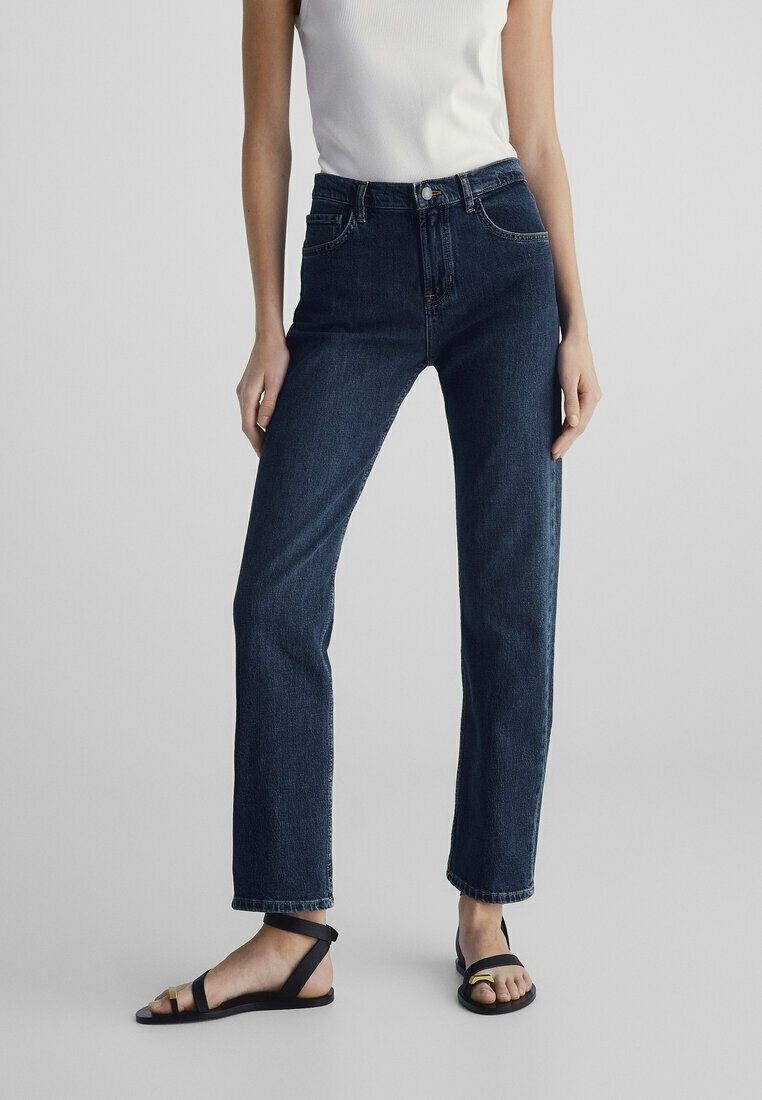 Femme portant un jean bleu foncé droit, un haut blanc sans manches et des sandales plates noires avec un détail doré sur un fond uni.