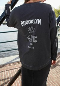 Musta pitkähihainen collegepaita, jossa on valkoista tekstiä, joka lukee "BROOKLYN 87 ESTABLISHED NYC." Vaatteessa on rento leikkaus ja pyöreä kaula-aukko.