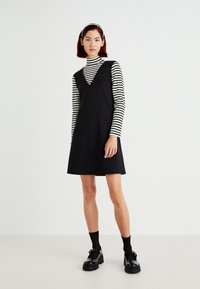 Vestido negro en corte A con escote en V, usado sobre una camiseta de manga larga a rayas blancas y negras. La modelo lleva zapatos negros gruesos y una diadema.