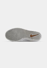 Podplat čevlja Nike ima teksturirano gumirano površino s satastim vzorcem, večinoma bele barve, s subtilnim rjavim logotipom.