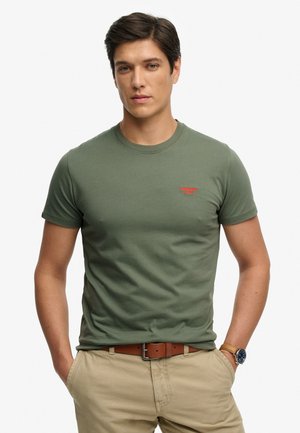 Ung mand iført olivengrøn t-shirt, beige bukser med brunt bælte, stående med hænderne i lommerne mod en ensartet lys baggrund.