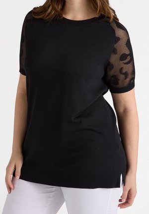 Vrouw draagt een zwart gebreid topje met transparante korte luipaardprintmouwen, gecombineerd met een witte broek tegen een effen achtergrond.