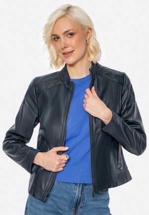 Blonde vrouw in een zwart leren jack over een blauwe sweater en jeans, licht glimlachend en de rits van het jack vasthoudend.