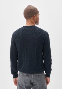 Mornarsko modra pletena jopica z teksturiranimi vzorci, z okroglo ovratnico in rebrastimi manšetami. Obuta z sivo jeans hlačami, videna s hrbtne strani.