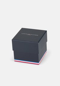 Boîte en papier noire avec un couvercle, ornée d'une bande rouge, blanche et bleue à la base et du logo "TOMMY HILFIGER WATCHES" sur le dessus.
