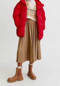 Červená péřová bunda, krémový svetr s barevnými vzory, béžová skládací sukně a hnědé robustní kotníkové boty s bílými ponožkami.