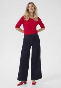 Punainen ribbipaita, jossa on aaltoilevat hihat, yhdistettynä korkeavyötäröisiin, leveisiin tummiin farkkuihin ja punaisiin paksupohjaisiin kenkiin.