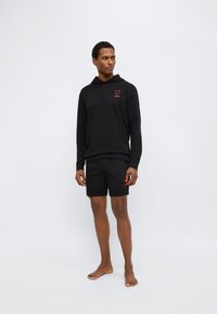 Μαύρο μακρυμάνικο φούτερ με κουκούλα και κόκκινο λογότυπο καρδιάς, συνοδευόμενο από μαύρο σορτς από το ίδιο λείο ύφασμα, φορώντας τα ξυπόλυτος σε ουδέτερο φόντο.