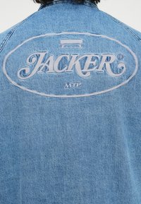 Svijetloplava traper jakna s velikim vezom "JACKER" logotipom na leđima, glatke teksture i vidljivim detaljima šivanja.