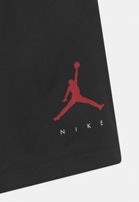 Červené logo basketbalového hráče Jumpman nad bílým textem "NIKE" na černé texturované tkanině, součást sportovního oděvu.