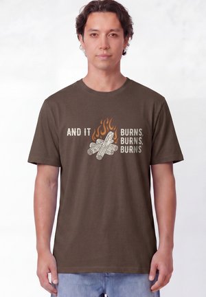 Junger Mann mit braunem T-Shirt mit Lagerfeuergrafik und dem Text "And it burns, burns, burns", stehend vor weißem Hintergrund.