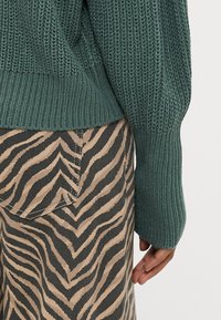 Πλεγμένο πράσινο πουλόβερ με ριμπ ύφασμα και μακριά μανίκια, συνδυασμένο με καφέ παντελόνι που διαθέτει μαύρο ζιγκ-ζαγκ μοτίβο και λεπτομέρεια πίσω τσέπης.