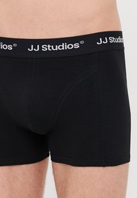Schwarze Boxer Shorts aus einem weichen Material, mit einem schwarzen Bund und dem weißen "JJ Studios"-Logo sowie dezenten Nähdetails.