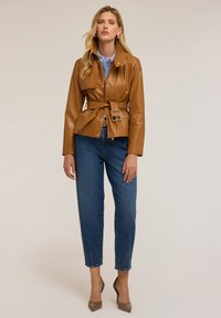 Veste en cuir marron avec ceinture, grand col, accessoires en métal argenté et poches zippées, associée à un jean bleu et des escarpins beiges.