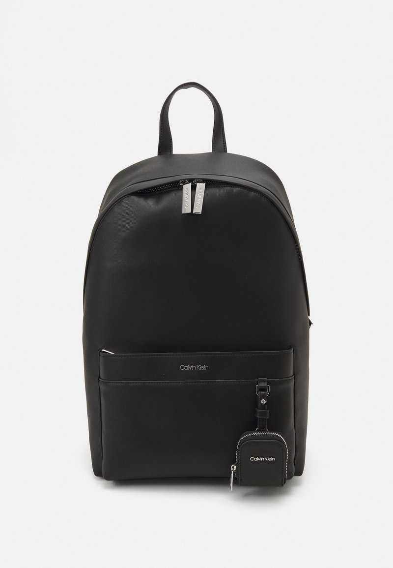 Black synthetic backpack with smooth texture, rounded shape, double zipper, front pocket, and attached small pouch. Calvin Klein logo visible.