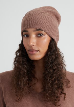 Vrouw met lang krullend haar, die een geribde bruine beanie en een bijpassende bruine V-hals trui draagt, tegen een effen achtergrond.