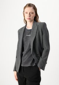 Grå rutete blazer med strukturerte skuldre, notched lapels og en enkelt knappelukking, båret over en mørk mønstret T-skjorte og svarte bukser.