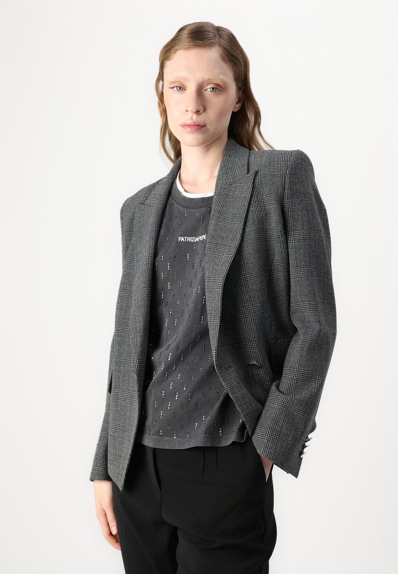 Szara marynarka w kratę z wyraźnymi ramionami, klapami w szpic i zapięciem na jeden guzik, noszona na ciemno wzorzystej koszulce oraz czarnych spodniach.