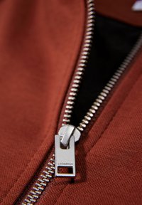 Rostfärgad zippered hoodie med en silverfärgad metizipper och en rektangulär dragflik. Tyget är slätt med en lätt struktur.