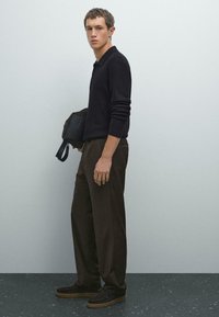 Jonge man in een zwart geribd poloshirt met lange mouwen, lichtbruine wijde broek en donkere instappers, terwijl hij een donkerbruine tas tegen een effen muur vasthoudt.