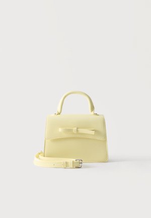 Piccola borsa giallo pallido con manico superiore, pattina frontale, fiocco decorativo e tracolla removibile posata su superficie bianca.