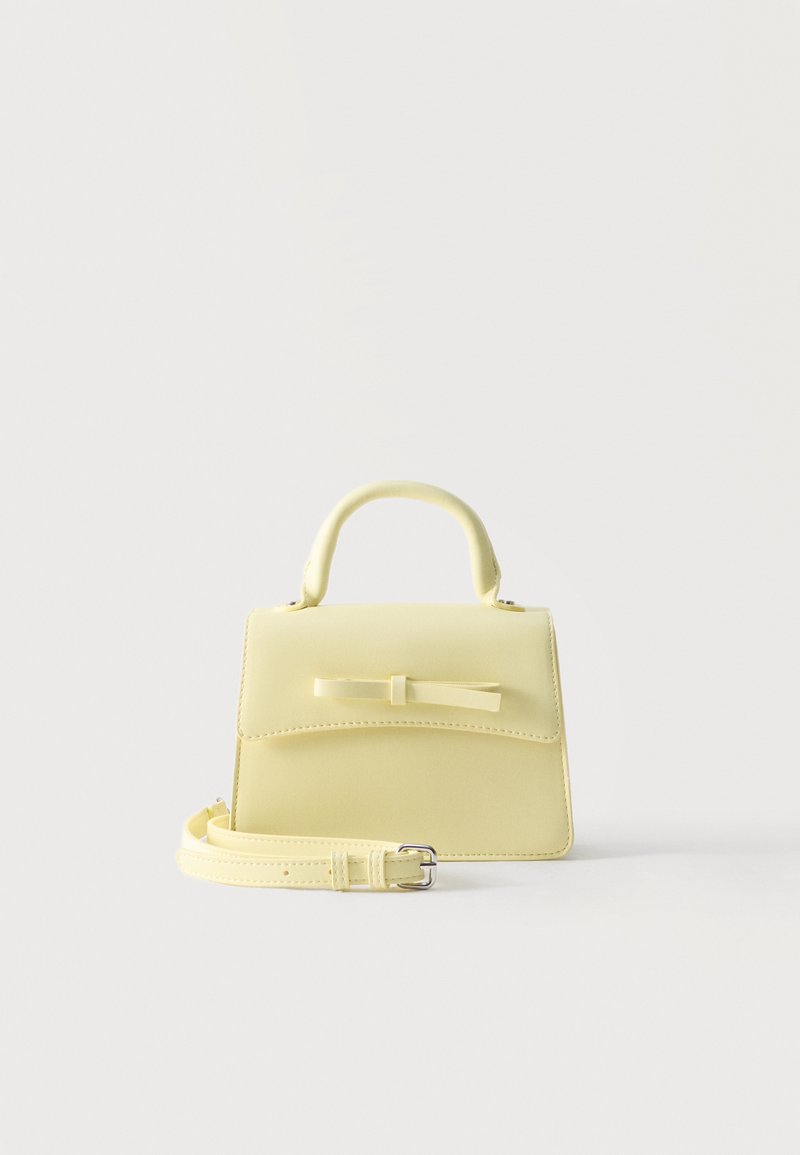 Petit sac à main jaune pale avec poignée supérieure, rabat frontal, nœud décoratif et bandoulière amovible posé sur une surface blanche.