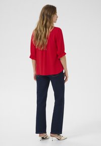 Blusa rossa a maniche corte con polsini arricciati, abbinata a jeans dritti blu scuro e décolleté beige a punta.