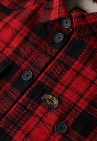 Czerwono-czarna flanelowa koszula w kratę z czarnymi guzikami i metką "Superdry". Tkanina ma miękką teksturę i przytulny wygląd.