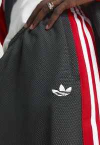 Svarta atletiska shorts med vit prickig struktur, med röda ränder och en vit broderad Adidas treklöverlogotyp på sidan.