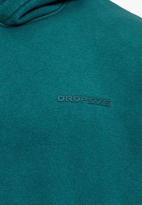 Gros plan sur un tissu bleu sarcelle avec le texte embossé subtil "DROPSIZE" centré sur une surface lisse et douce au toucher.