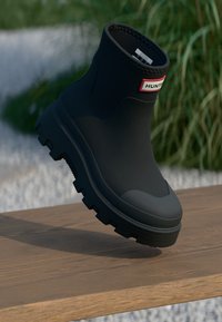 Μαύρο ελαστικό μποτάκι μέχρι τον αστράγαλο με υφασμάτινη σόλα, φέρει ετικέτα "HUNTER" σε κόκκινο και λευκό χρώμα. Λείο σχέδιο και επενδεδυμένο άκρο.