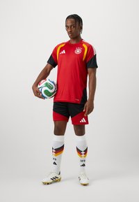 Κόκκινη και μαύρη αθλητική φανέλα με πλέγμα υφής, σε συνδυασμό με μαύρα σορτς. Λευκές κάλτσες με διαγώνιο χρωματικό σχέδιο. Ο παίκτης κρατάει μια μπάλα ποδοσφαίρου.