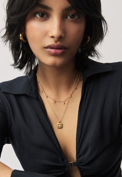 Arany bevonatú nyakláncok négyzet alakú medálal és kis díszgömbökkel, fekete gombos inggel viselve, bemutatva egy puha szövet textúrát.