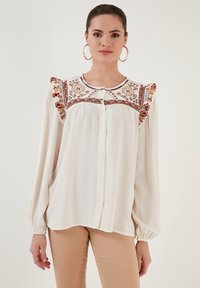 White long-sleeve blouse with decorative embroidered neckline and ruffle accents. Features button front and soft, lightweight fabric.