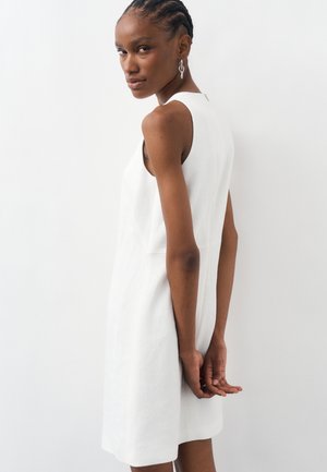 Naine palmikutega juustega, kandes valget varrukateta kleiti ja hõbedast kettidest kõrvarõngaid, seisab külje peal, käed selja taga kokku pandud.