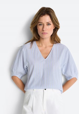 Žena s vlasmi po ramená v modro-bielej blúzke s vertikálnymi pruhmi a nafúknutými rukávmi, oblečená v bielych nohaviciach, stojaca na bielom pozadí.