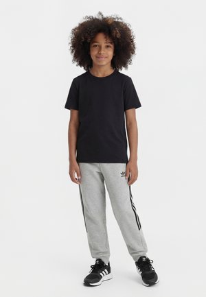 Niño con cabello rizado que está de pie vistiendo una camiseta negra, pantalones deportivos grises de Adidas y zapatillas negras Adidas sobre un fondo blanco liso.
