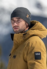 Žlutá nepromokavá bunda s černou nášivkou s logem "HH" na rukávu. Šedá pletená čepice. Sněhové pozadí.