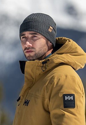Žlutá nepromokavá bunda s černou nášivkou s logem "HH" na rukávu. Šedá pletená čepice. Sněhové pozadí.
