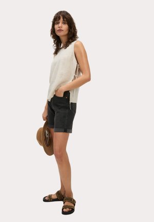 Un haut sans manches en ribbed beige clair avec des liens sur le côté, assorti à un short en denim foncé et des sandales cloutées. Le modèle tient un chapeau en paille marron.