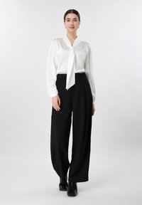 Jeune femme debout, portant un chemisier blanc avec cravate et un pantalon large noir avec des bottines noires, fond neutre.