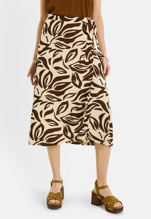 Mujer con falda midi beige cruzada con estampados abstractos de hojas en marrón oscuro y sandalias de plataforma marrones con tiras en el tobillo.