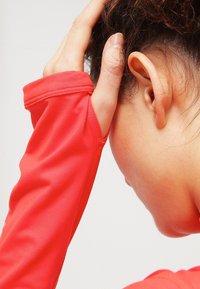 Henkilö, jolla on kihara hiukset, pitää päätään kädellä punaisessa pitkähihassa paidassa, näkyvät korvat ja sivuvaltimo yksinkertaista taustaa vasten.
