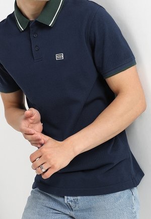 Homme portant un polo Levi's bleu marine avec un col bordé de vert et un jean bleu clair, les mains jointes devant lui.