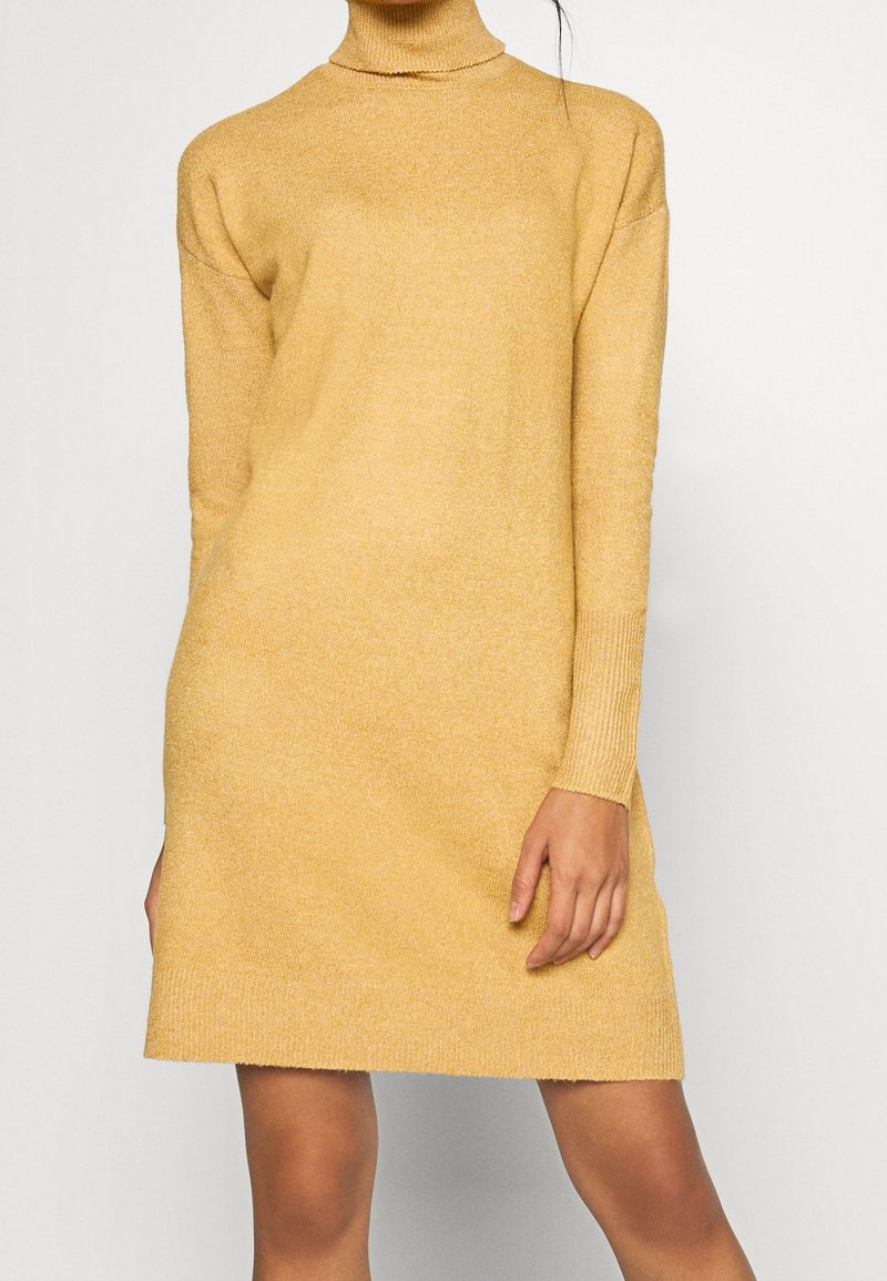 Femme portant une robe en maille jaune moutarde à col roulé et manches longues, debout devant un fond blanc uni.